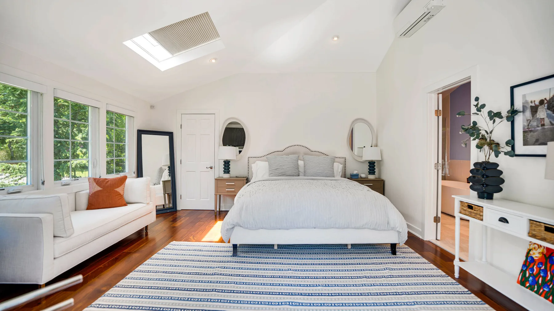Un dormitorio luminoso con una cama tendida, asientos blancos y ventanales.