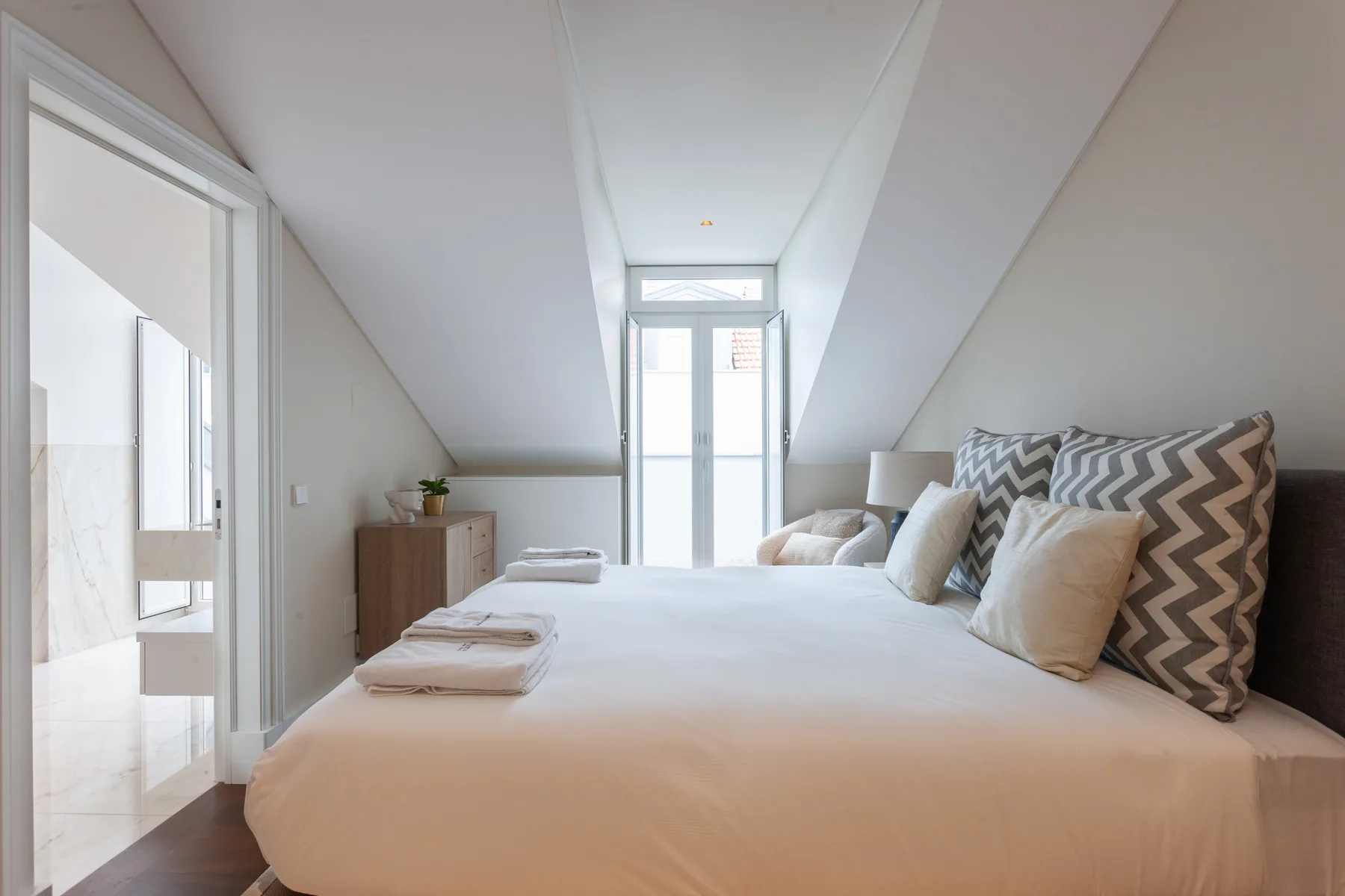 A bright bedroom with a bed beneath a sloped ceiling and tall windows.