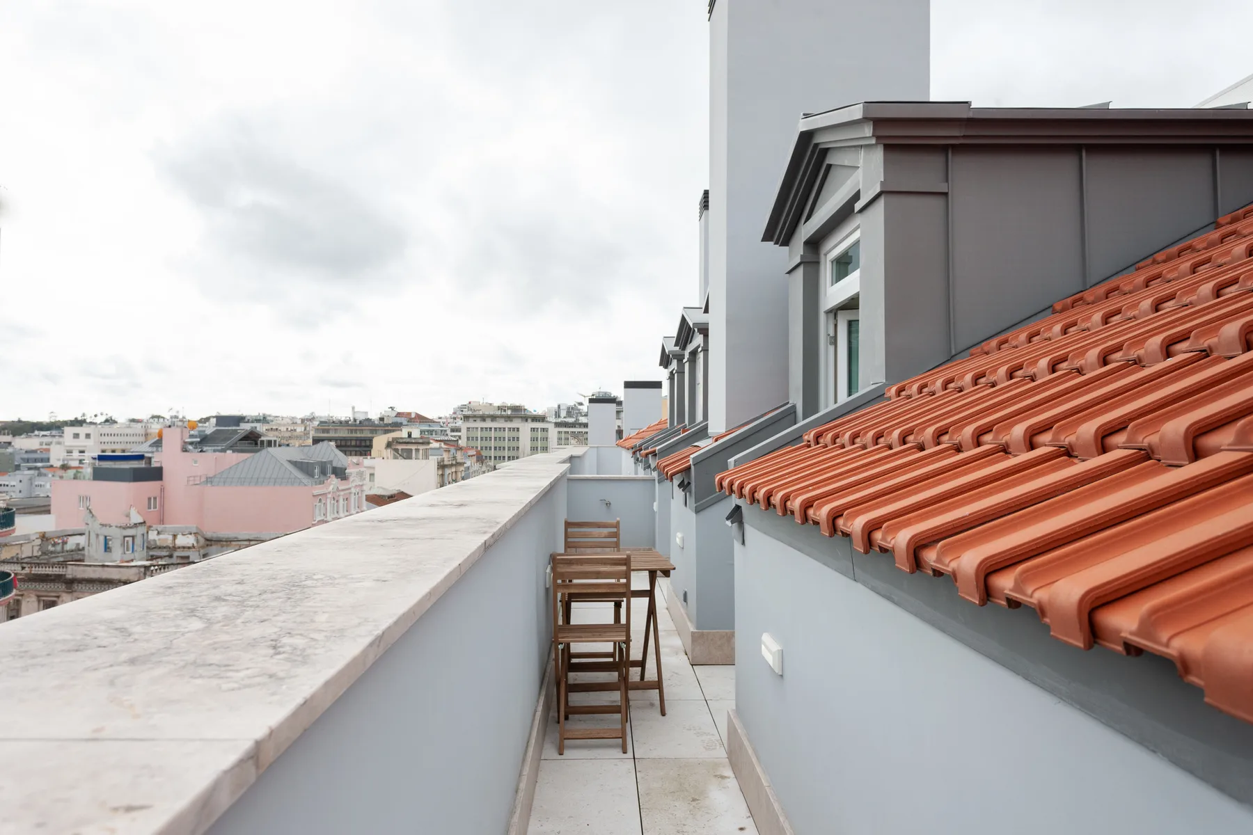 A narrow rooftop terrace with a stone parapet, stools, and views of nearby buildings.