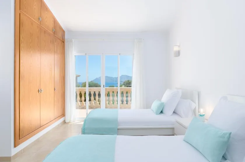 Un dormitorio luminoso con dos camas y un armario de madera junto a una ventana con balcón.