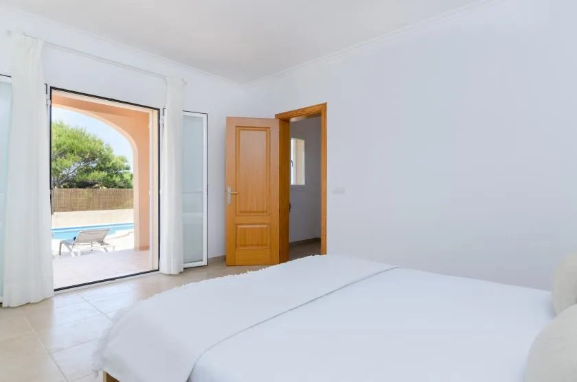 Un dormitorio luminoso con una cama junto a puertas abiertas que dan a una terraza con piscina.
