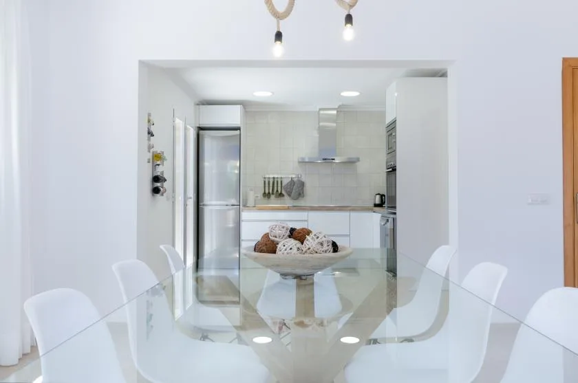 Un comedor luminoso con una mesa de vidrio y una cocina abierta al fondo.