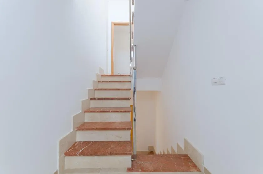 Una escalera con peldaños marrón rojizo sube junto a paredes blancas.