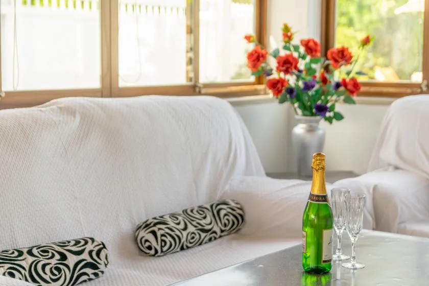 A bright sitting room with covered seating, flowers, and a bottle on a table.
