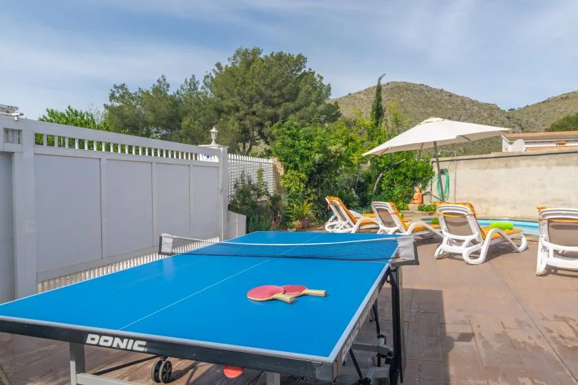 An outdoor table tennis table sits beside a swimming pool and sun loungers.