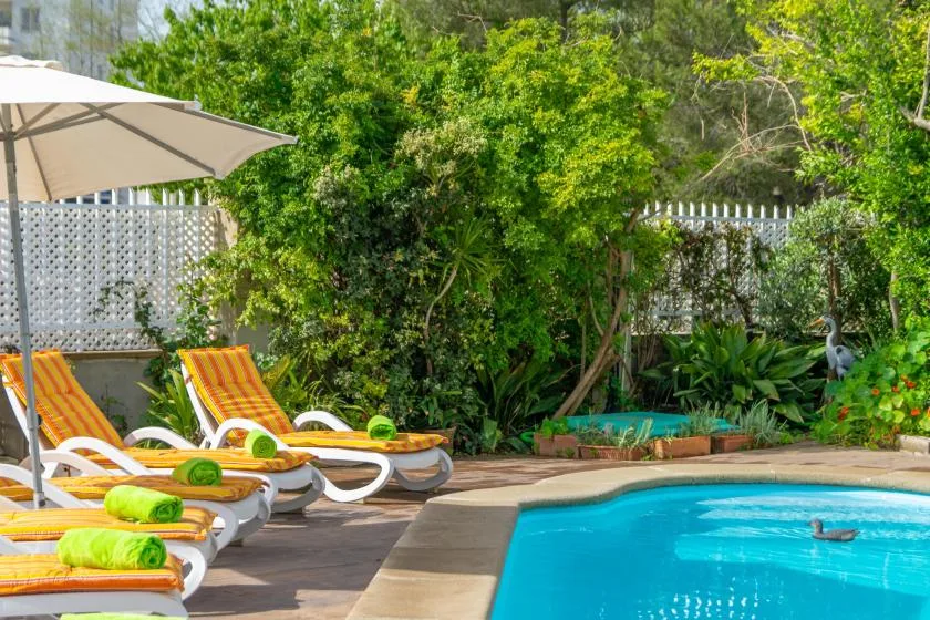 An outdoor swimming pool beside lounge chairs, an umbrella, and lush greenery.