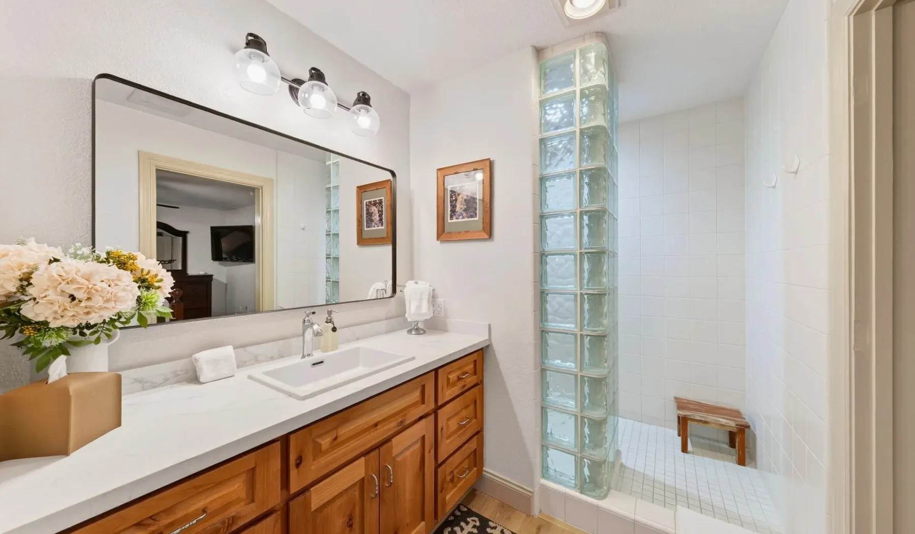 Un tocador de baño con un espejo grande está junto a una pared de bloques de vidrio de la ducha.