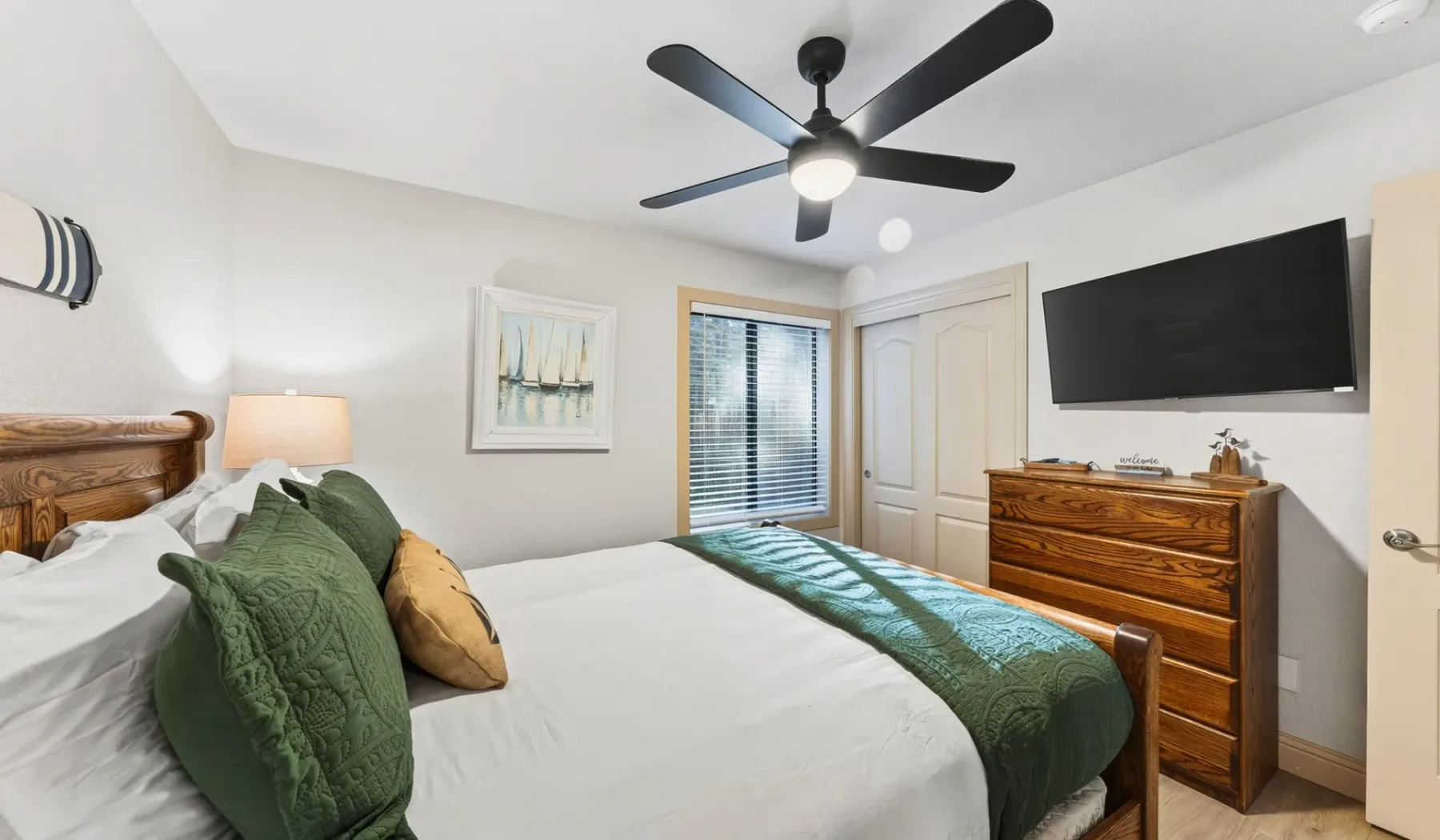 Un dormitorio con una cama tendida, ropa de cama verde y una cómoda bajo un televisor montado en la pared.