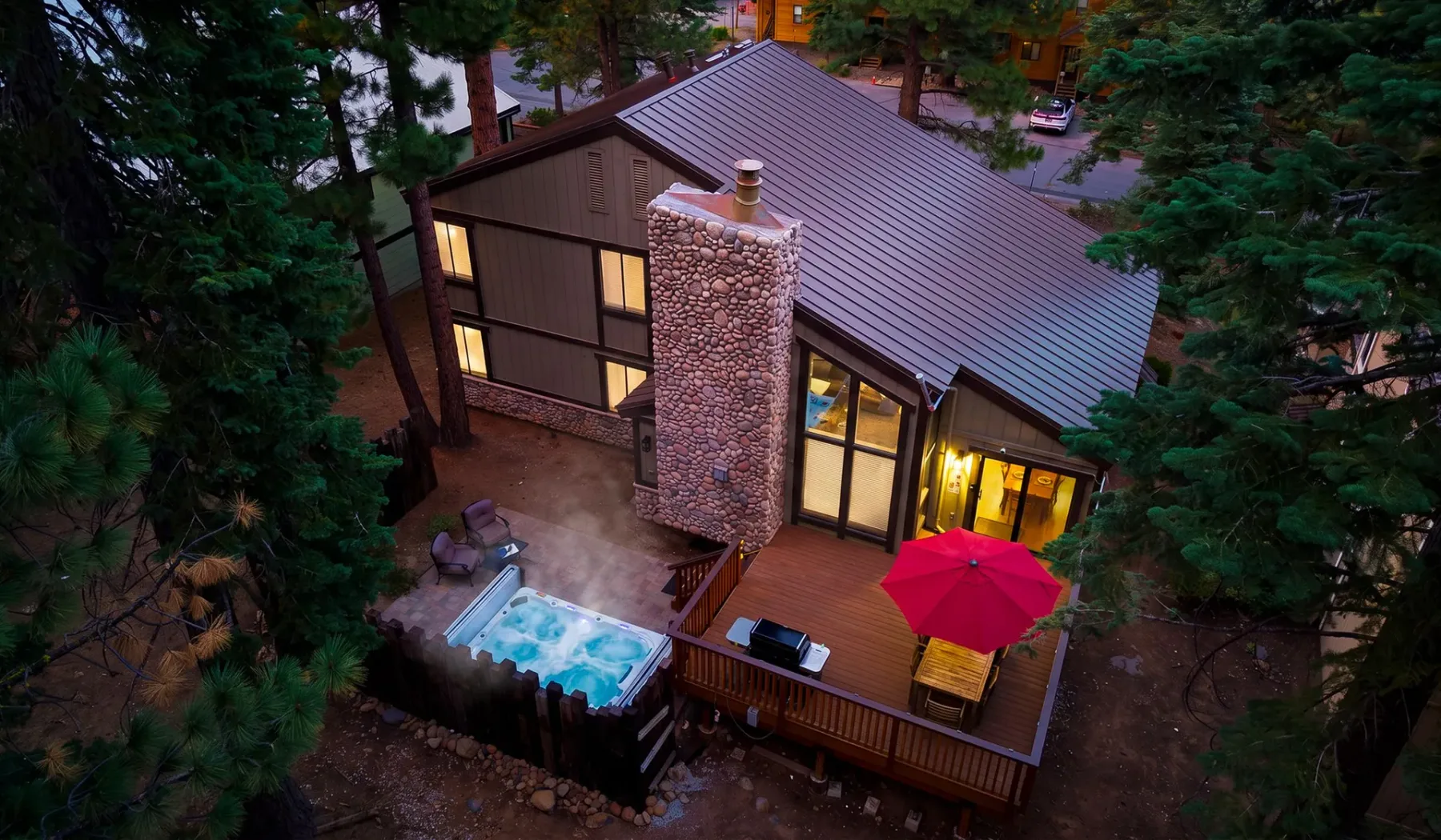 Una cabaña entre árboles tiene una chimenea de piedra, una terraza de madera y una bañera de hidromasaje exterior.
