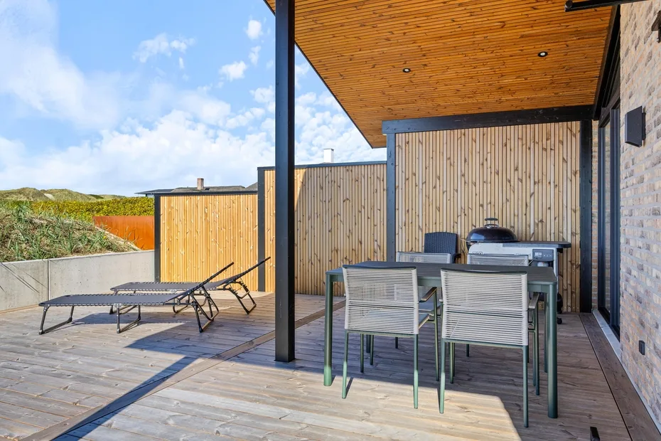 Comedor exterior cubierto con mesa, sillas y parrilla sobre una terraza de madera.