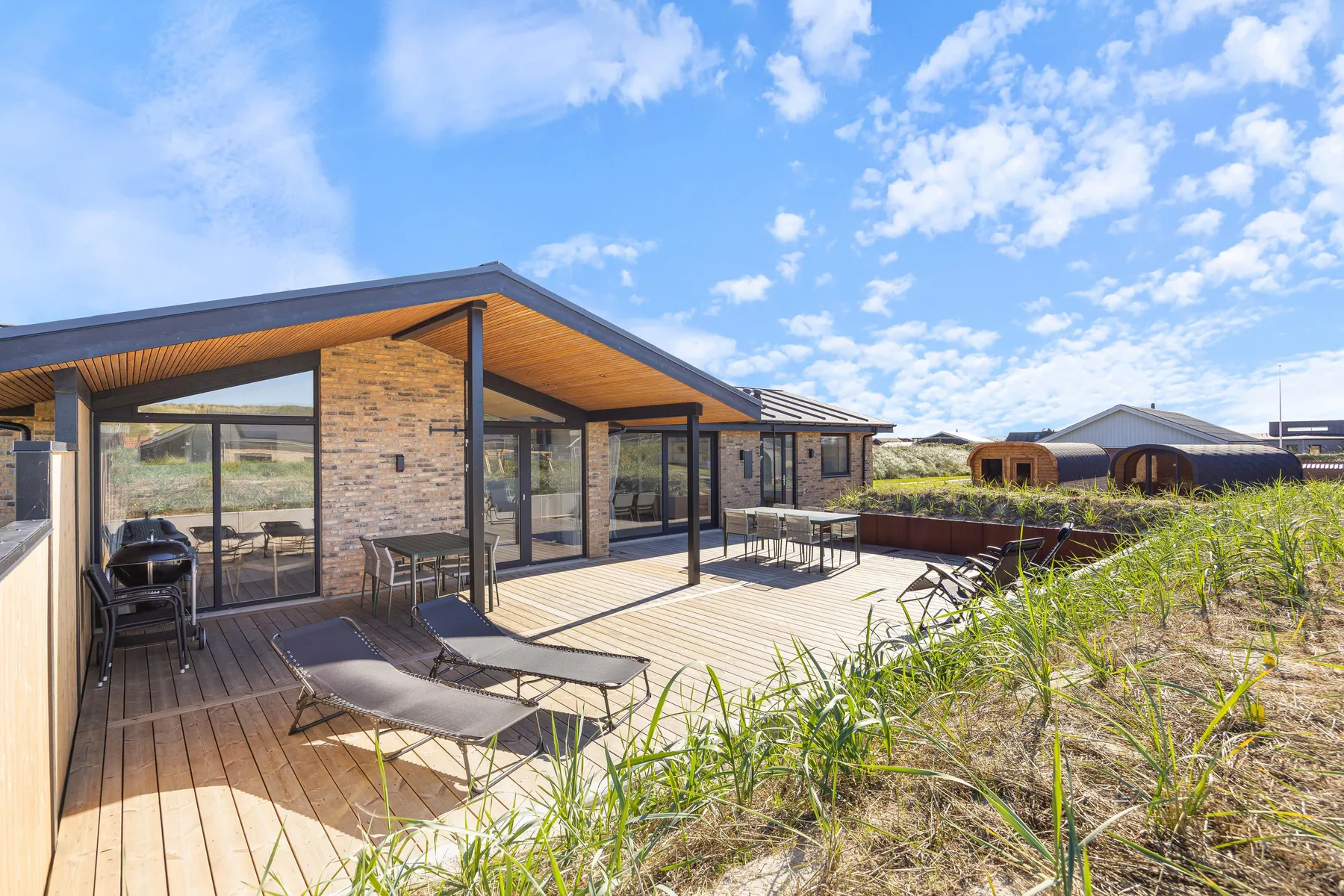 Una casa moderna se abre a una terraza de madera con asientos al aire libre y hierba cerca.