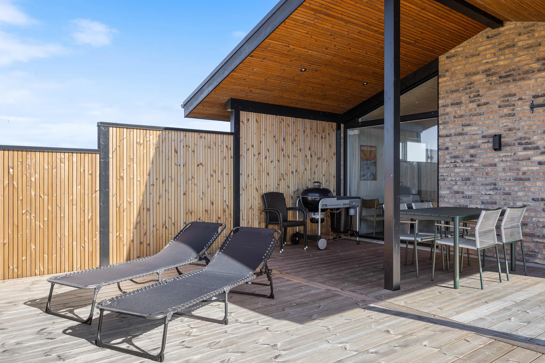 Una terraza de madera junto a una casa moderna tiene tumbonas y una zona de comedor exterior.