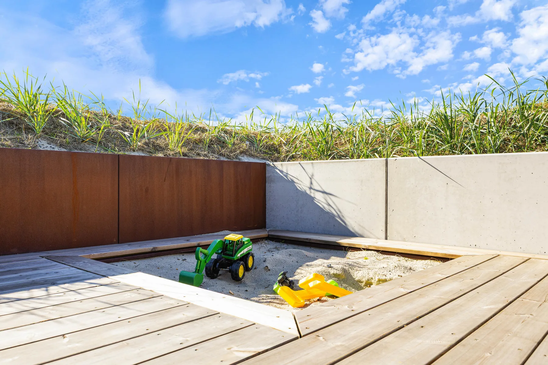 Una terraza de madera rodea una pequeña zona de arena con vehículos de juguete.