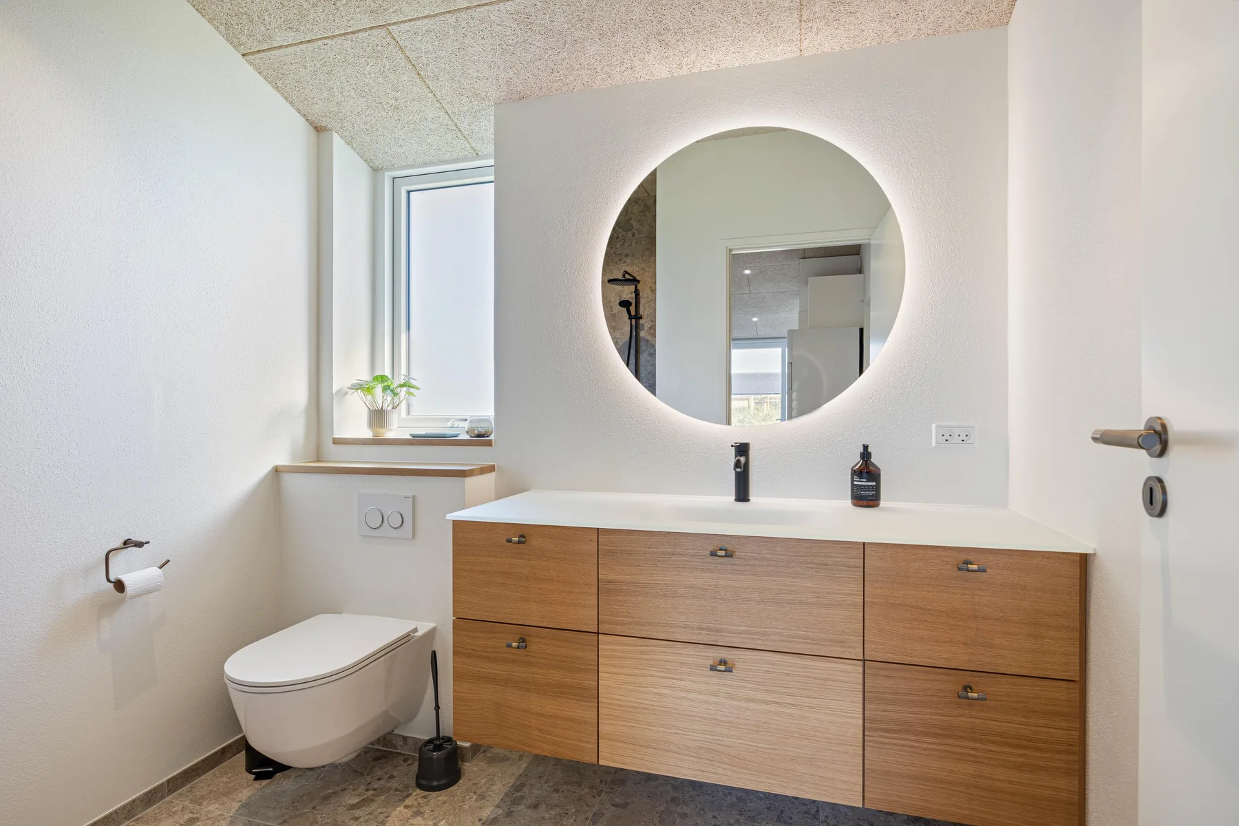 Baño luminoso con un mueble de madera y un espejo redondo sobre el lavabo.