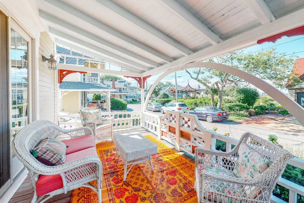 A covered porch with wicker seating and a colorful rug overlooks a parking area.