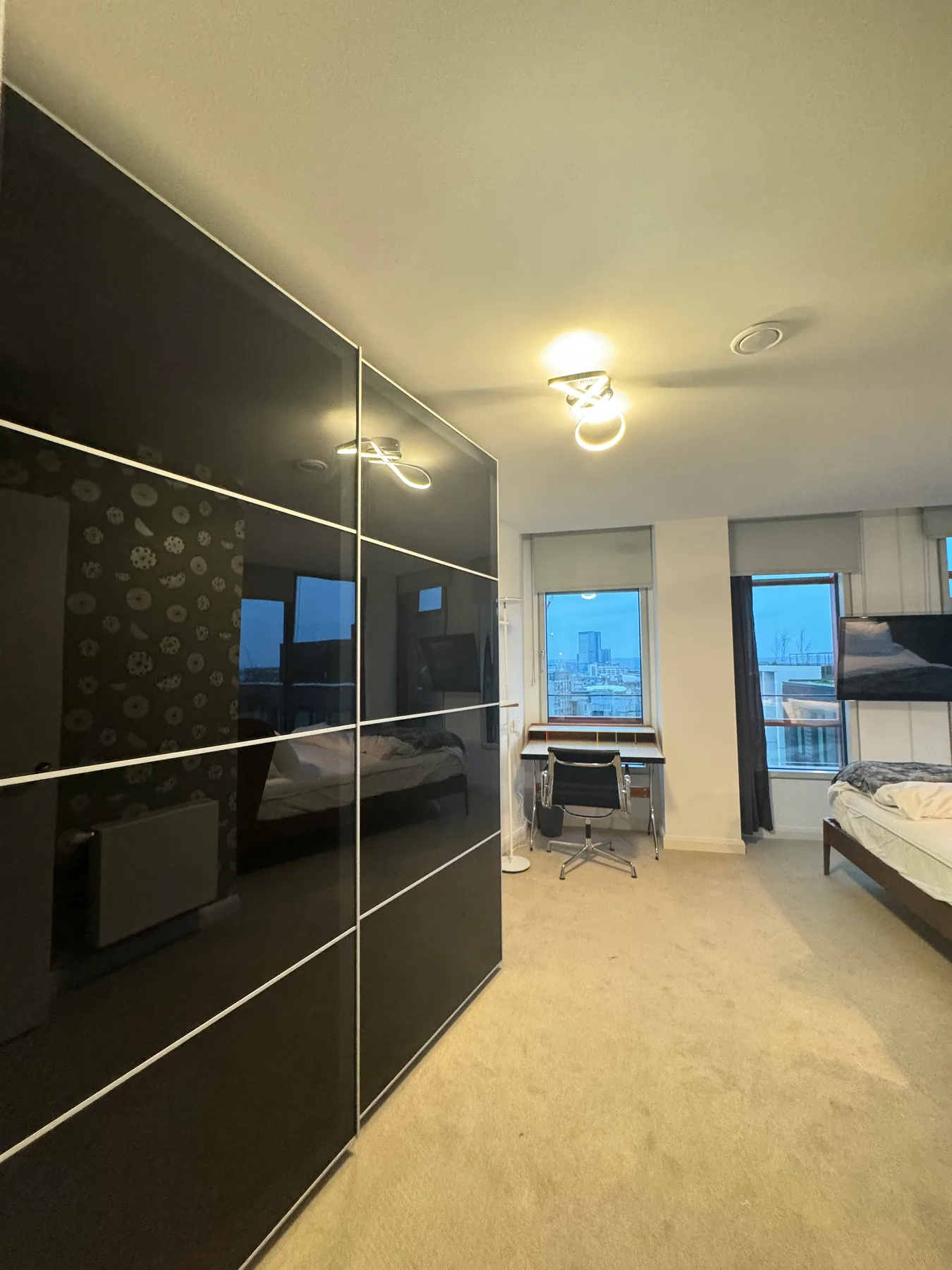 A bedroom with a bed, desk, and large windows overlooking the city.