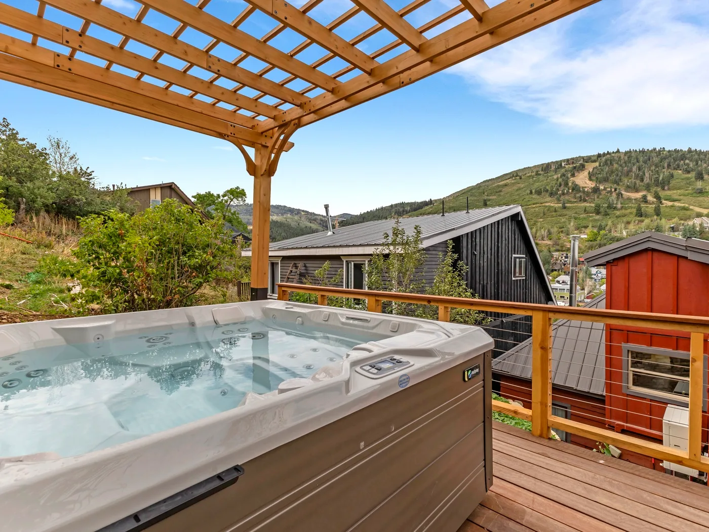 An outdoor hot tub sits on a wooden deck beneath a pergola, overlooking a hillside.