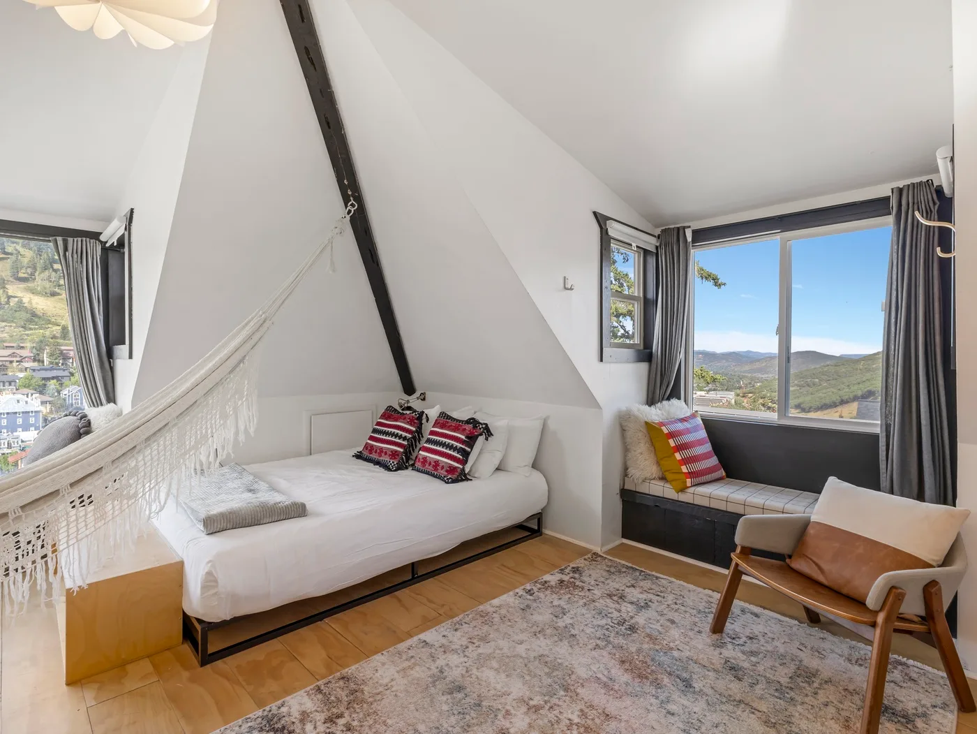 Un dormitorio abuhardillado luminoso con cama, hamaca y grandes ventanas con vistas a las colinas.
