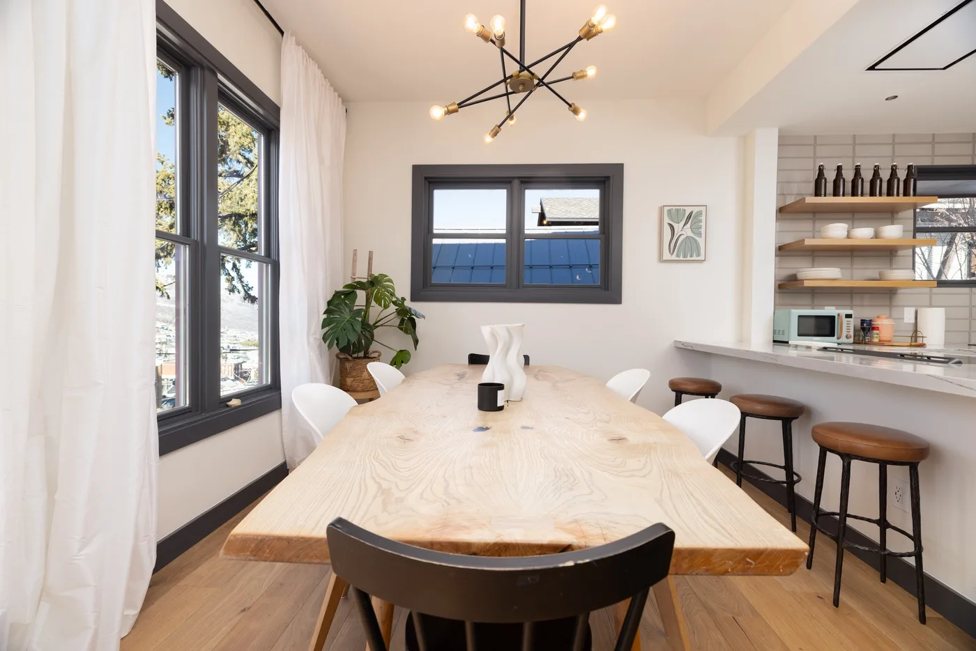 Un comedor luminoso con una mesa larga junto a grandes ventanas y una cocina abierta.