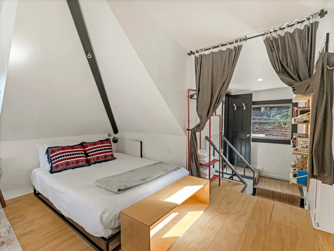 Un dormitorio con una cama baja bajo un techo inclinado y cortinas que dan a una escalera.