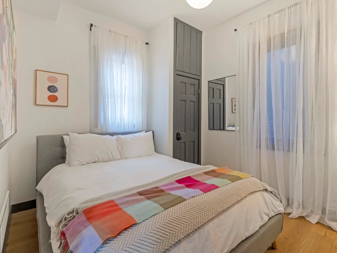 Un dormitorio con una cama, una manta colorida y ventanas altas con cortinas.