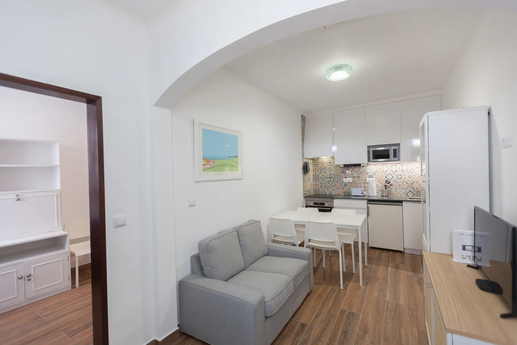 Una sala de estar comunica con una cocina y tiene un sofá gris y una mesa de comedor.