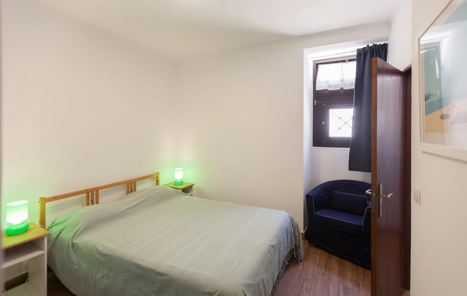 Un dormitorio con una cama bien tendida y un sillón oscuro junto a la ventana.