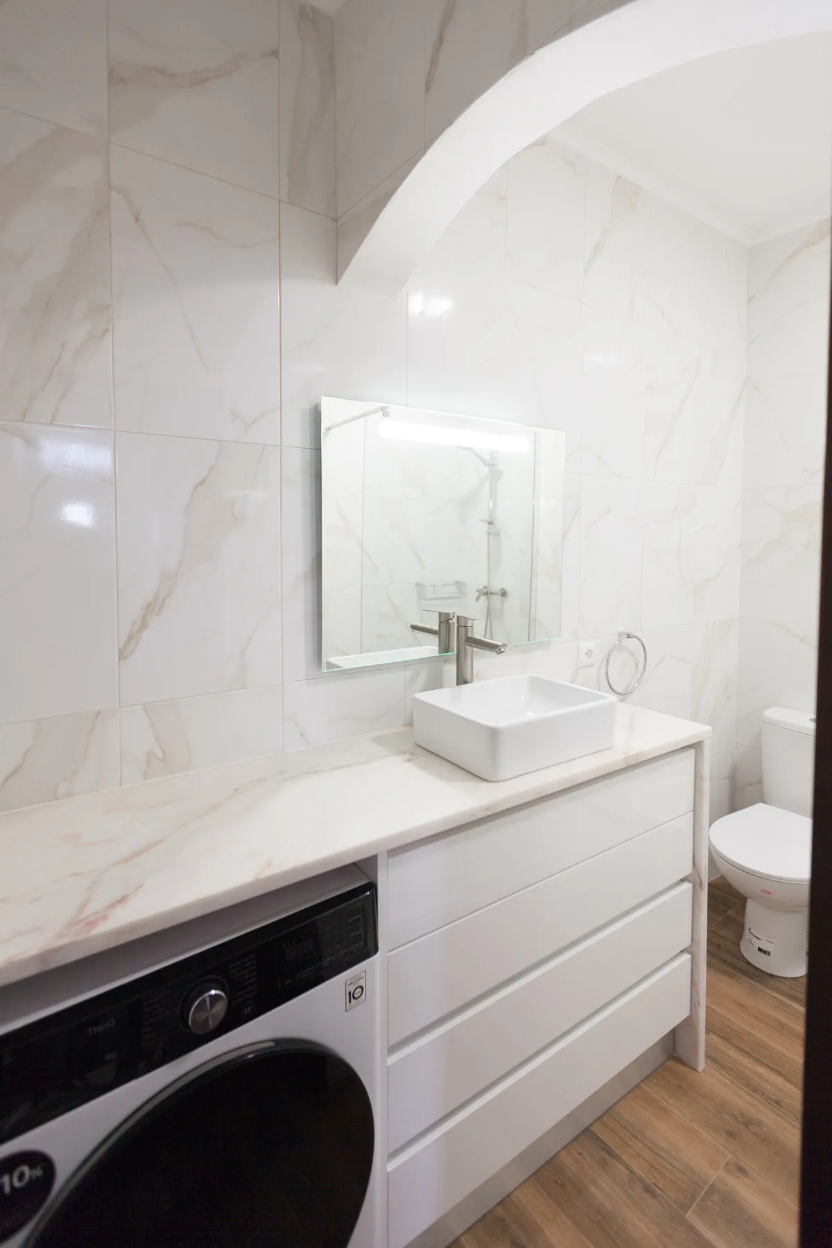 Baño luminoso con lavabo blanco, pared con espejo y lavadora.
