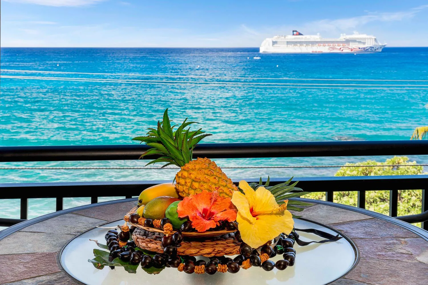 Un arreglo de frutas está sobre una mesa al aire libre, frente al agua turquesa y un crucero.