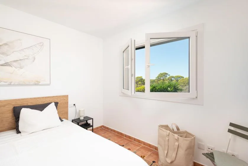 A bright bedroom with a bed beside an open window overlooking greenery.