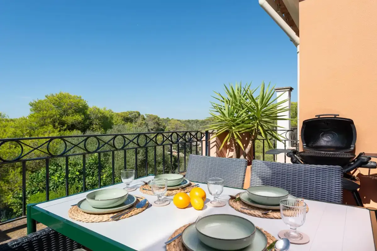 An outdoor dining table sits beside a terrace railing, with greenery beyond.