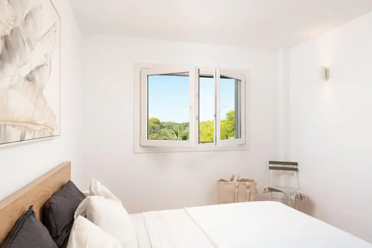 A bright bedroom with a bed beside an open window overlooking greenery.