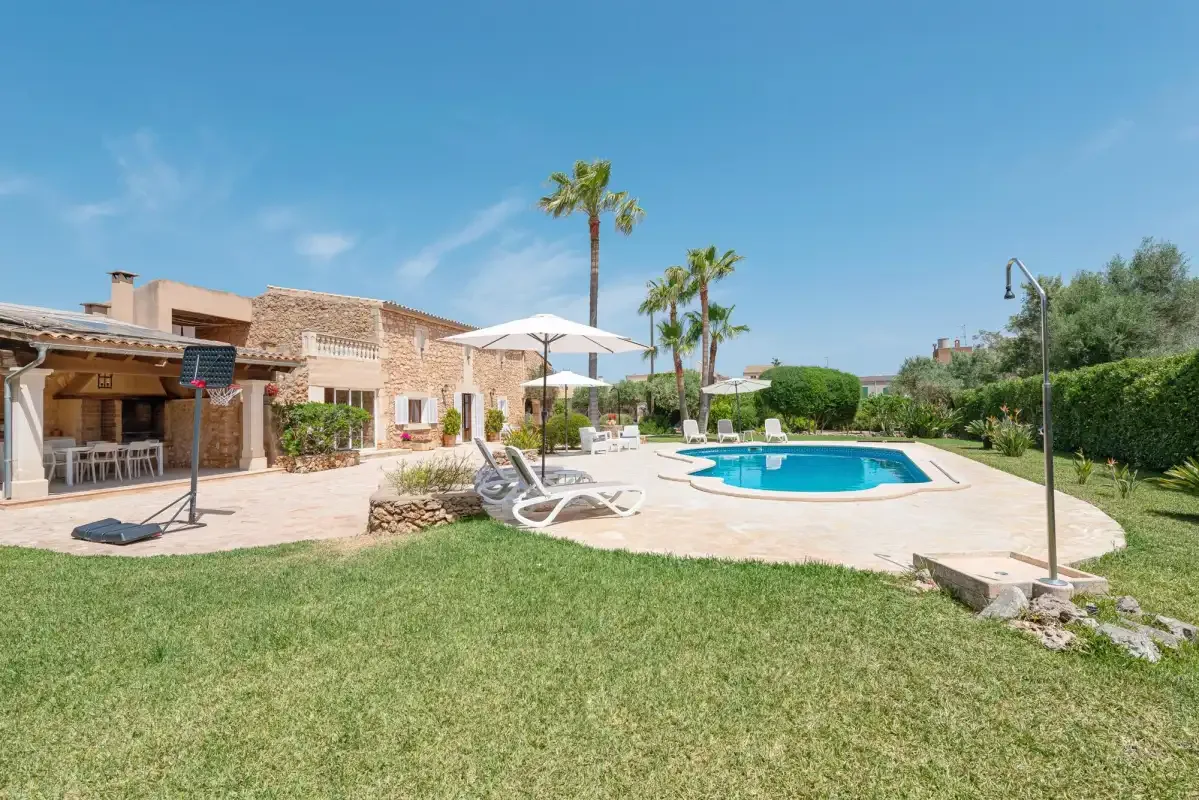 An outdoor swimming pool beside a stone house and lawn under a clear sky.