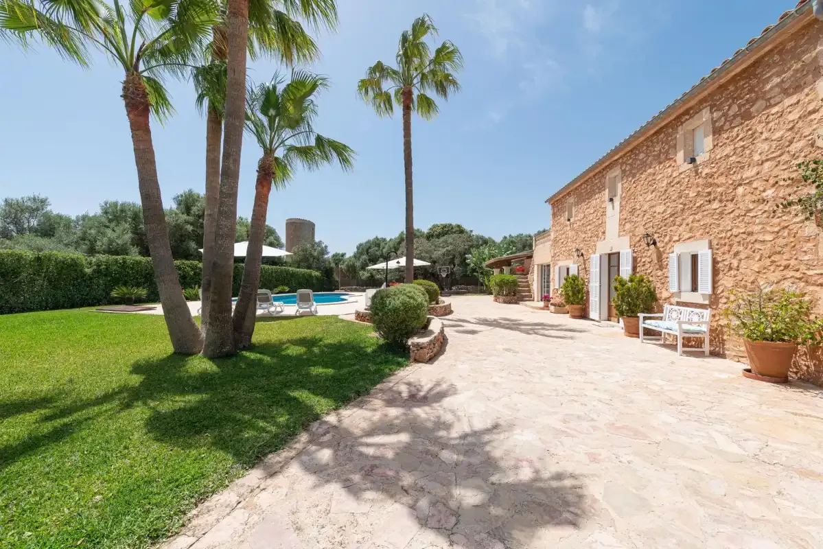 A stone house opens onto a sunny garden patio with palm trees and a pool.