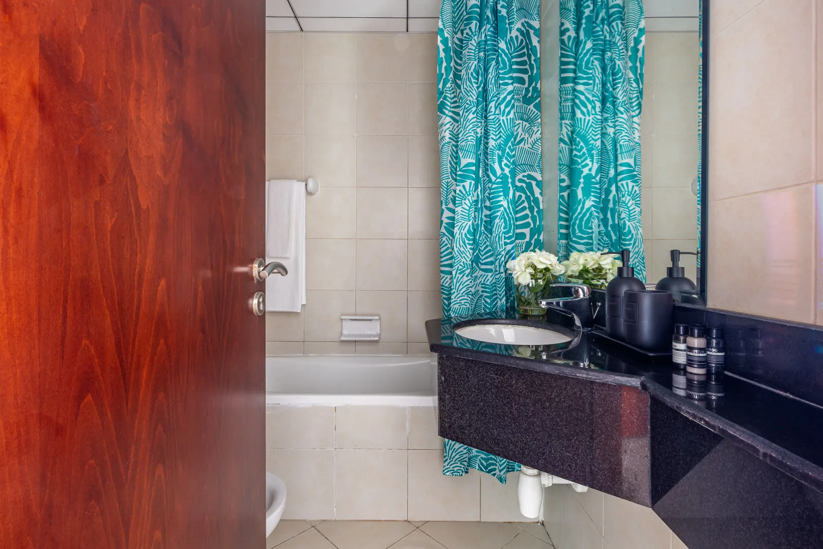 Un baño con bañera, paredes alicatadas y un tocador oscuro bajo una cortina estampada.