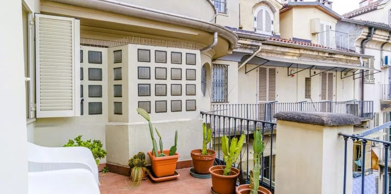 Un balcón con plantas en macetas, asientos al aire libre y fachadas de edificios cercanos.