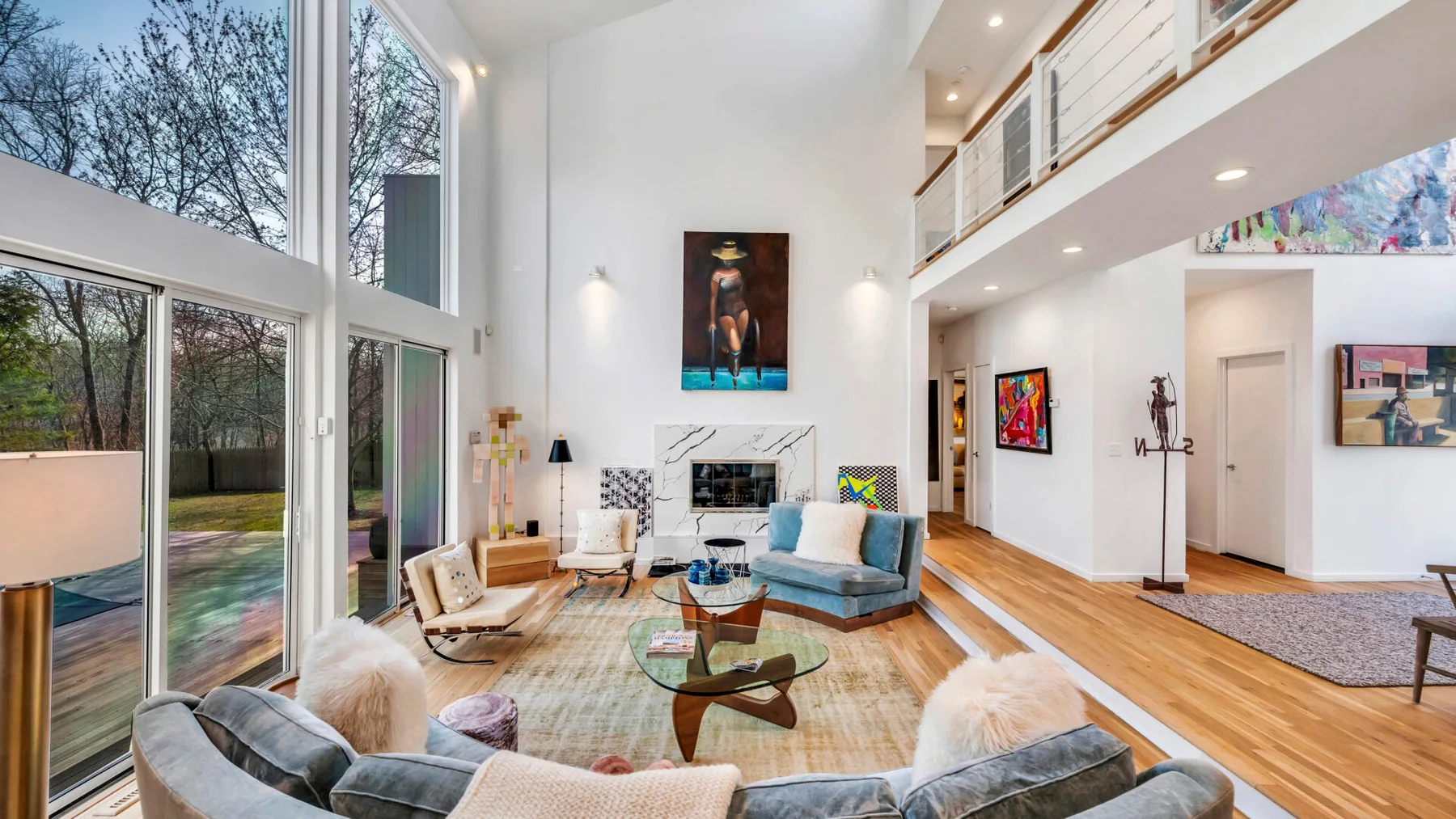 A bright double-height living room has large windows, seating, and a glass coffee table.