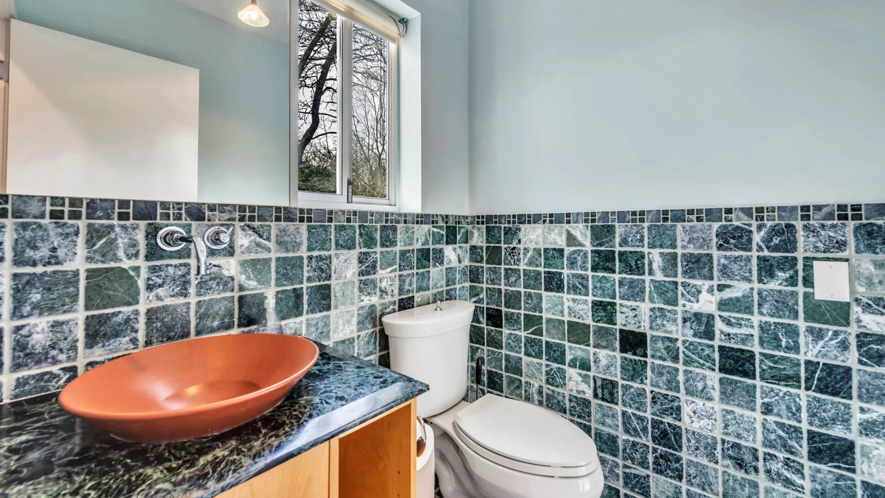 A bathroom with a dark tiled wall and a sink beside the toilet.