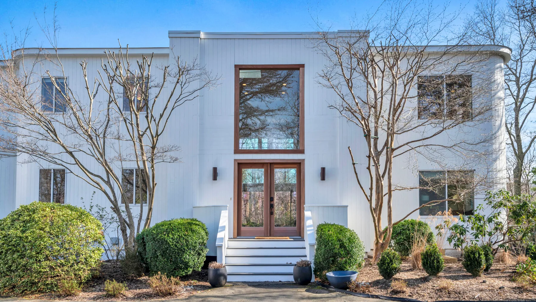 A white two-story house stands behind leafless trees and garden shrubs.