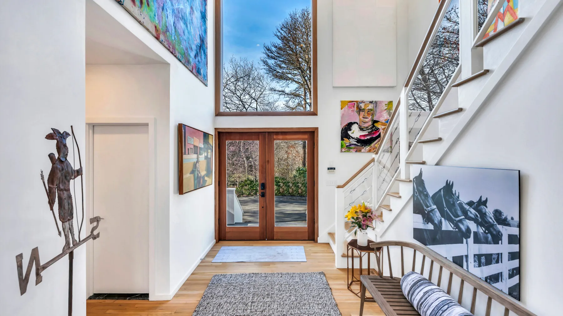 A bright double-height entry hall with a staircase, artwork, and glass doors.
