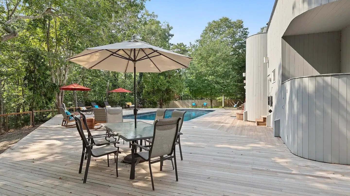 Una terraza junto a la piscina con una mesa, asientos de patio y una sombrilla grande.