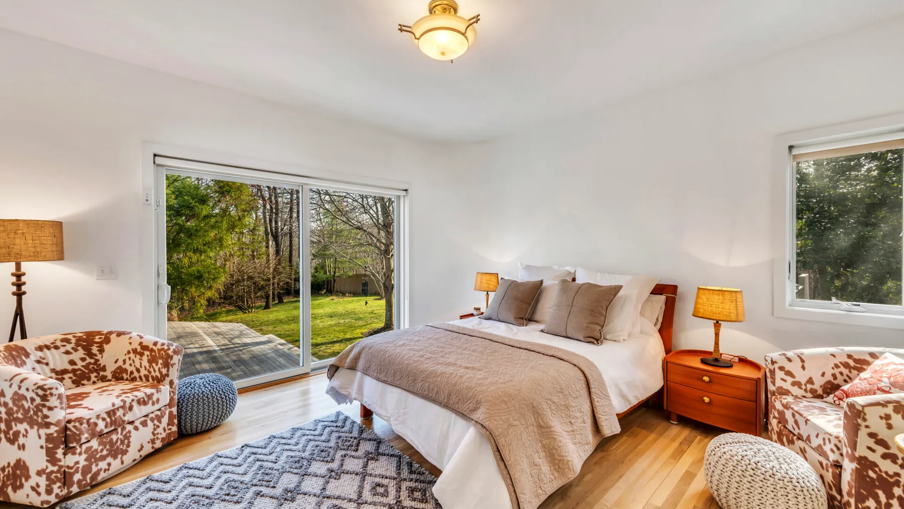 A bright bedroom with a bed, patterned seating, and large windows overlooking greenery.