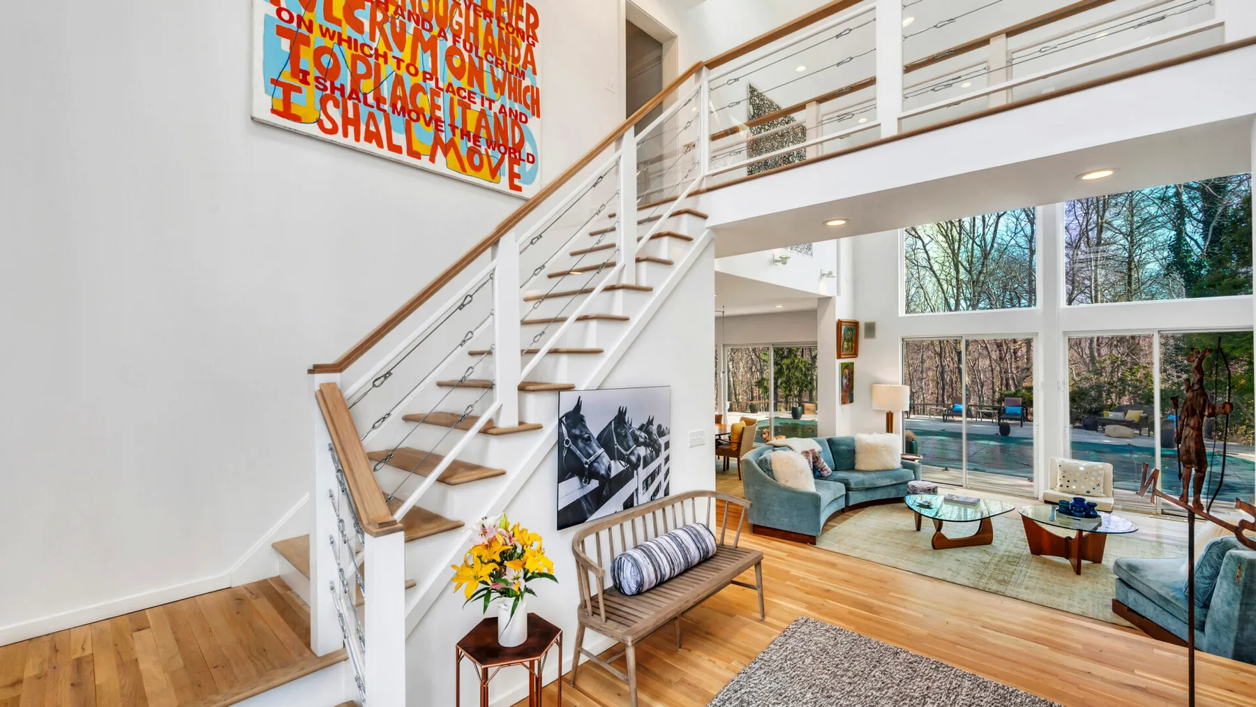 A bright living room with a staircase, seating, and large windows overlooking trees.