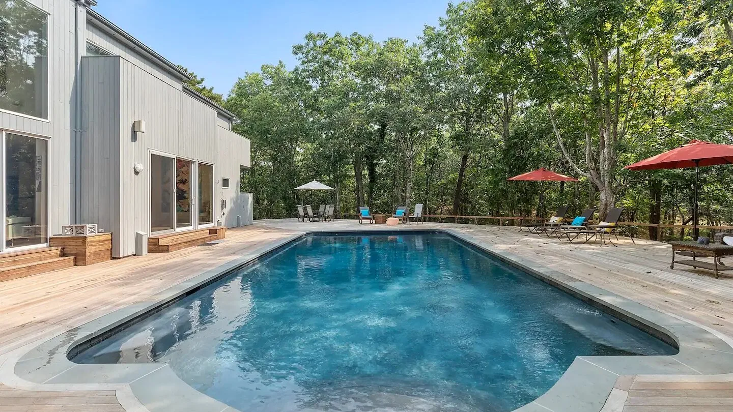 Una piscina al aire libre junto a una casa y una terraza rodeada de árboles.