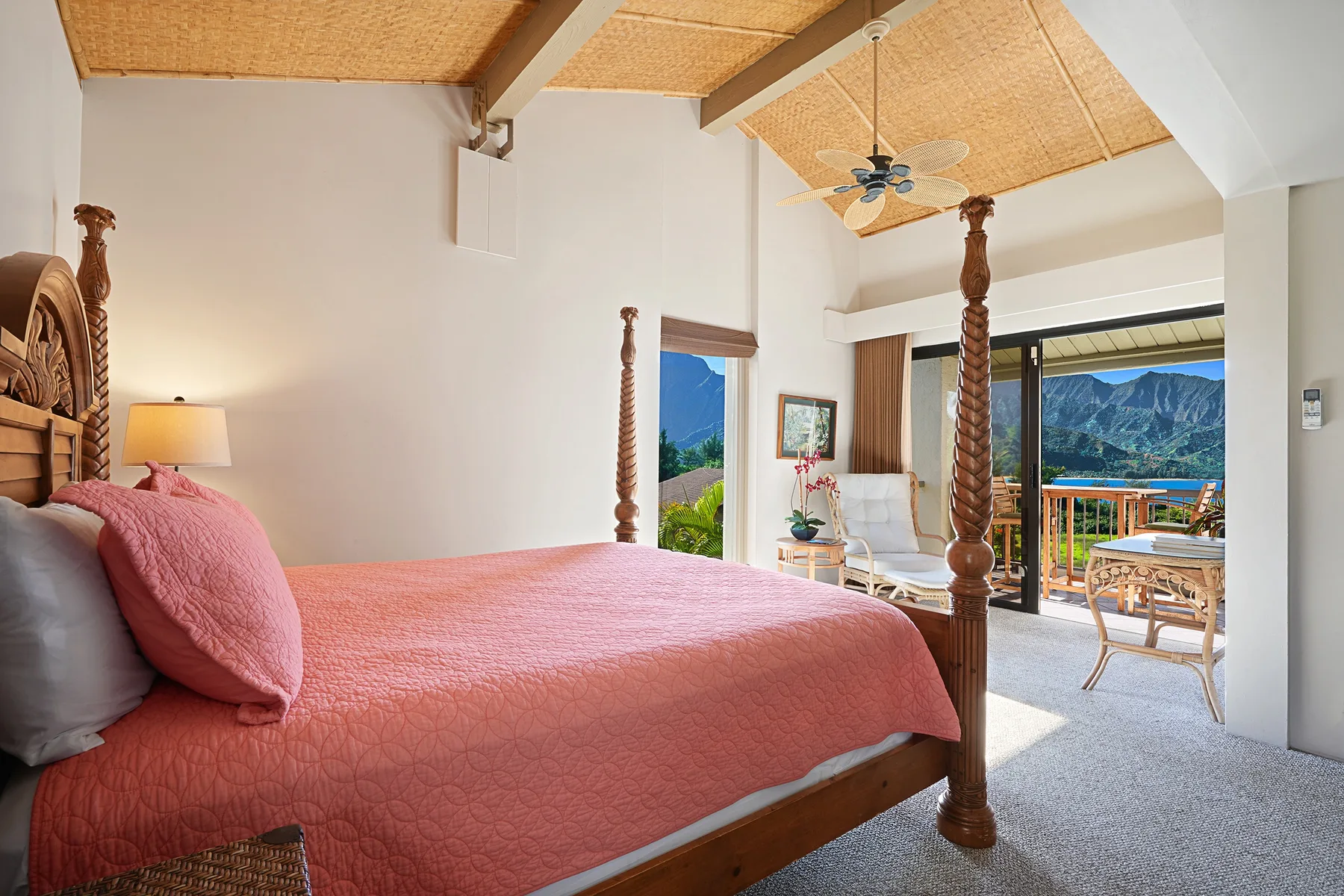Un dormitorio con una colcha rosa, ventilador de techo y puertas de vidrio hacia un balcón.