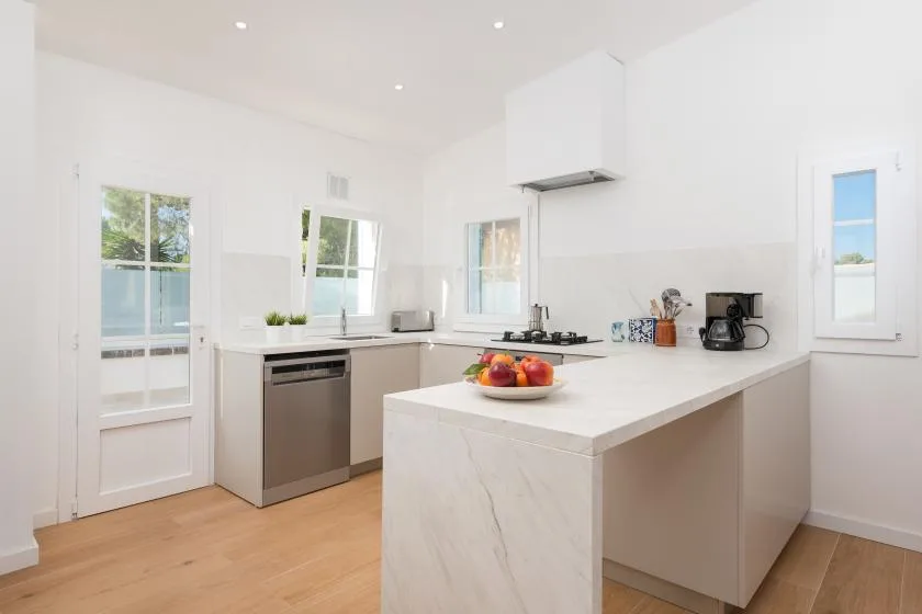 Una cocina blanca y luminosa con una isla central y suelo de madera clara.