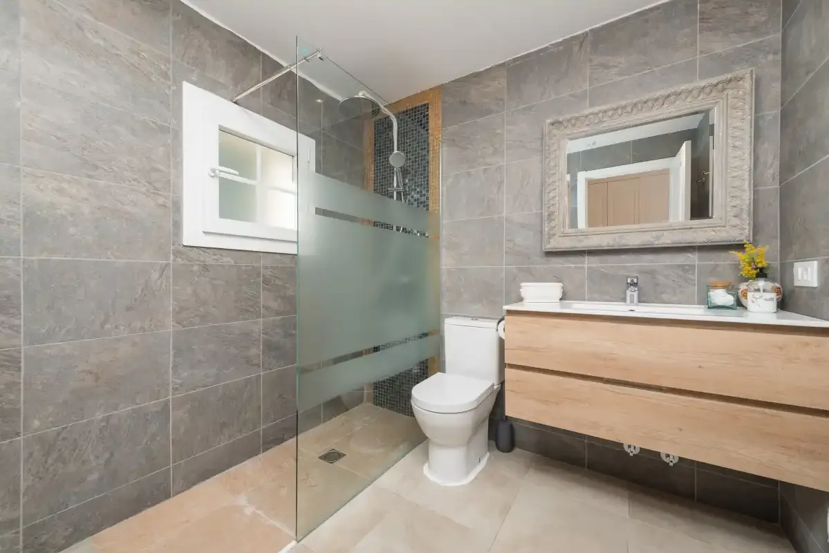 Un baño alicatado con ducha de vidrio, inodoro y mueble de lavabo en tonos de madera.