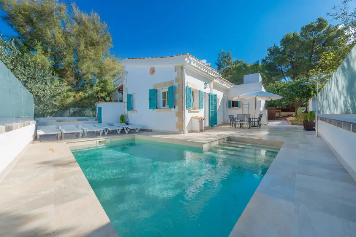 Una piscina y un patio pavimentado junto a una casa blanca con contraventanas turquesas.