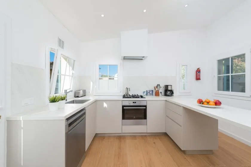 Una cocina moderna y luminosa con encimeras blancas y suelo de madera clara.