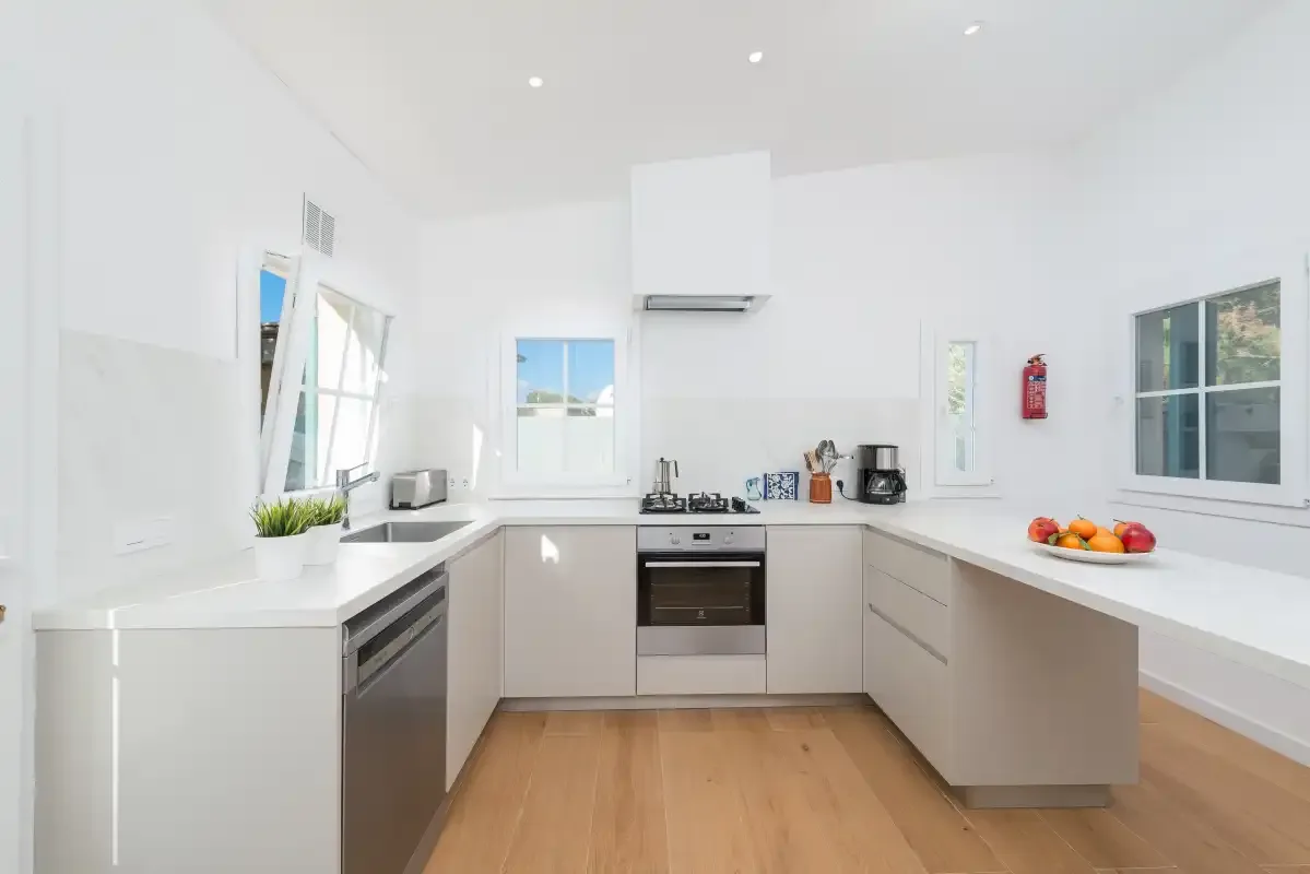Una cocina luminosa con encimeras blancas, gabinetes claros y suelo de madera.
